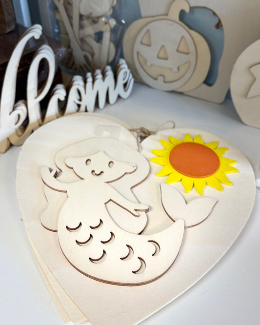 Wooden cutout of a mermaid with a sunflower on a heart-shaped board, surrounded by Halloween-themed decorations.