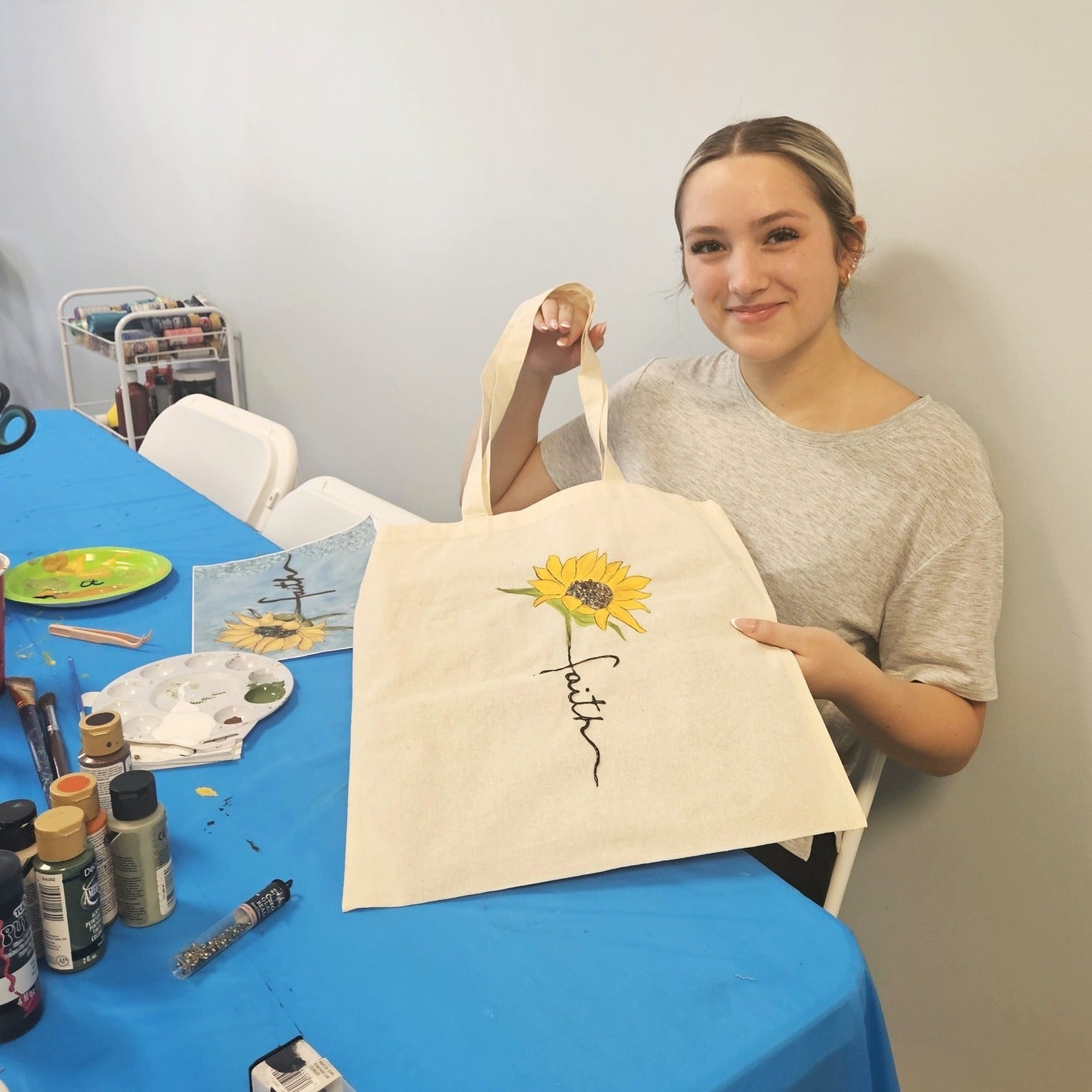 Woman holding a tote bag with a sunflower design and 'faith' text, standing in front of a table with art supplies.