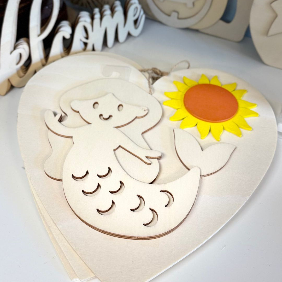 Wooden cutout of a mermaid with a sunflower on a heart-shaped board, surrounded by Halloween-themed decorations.