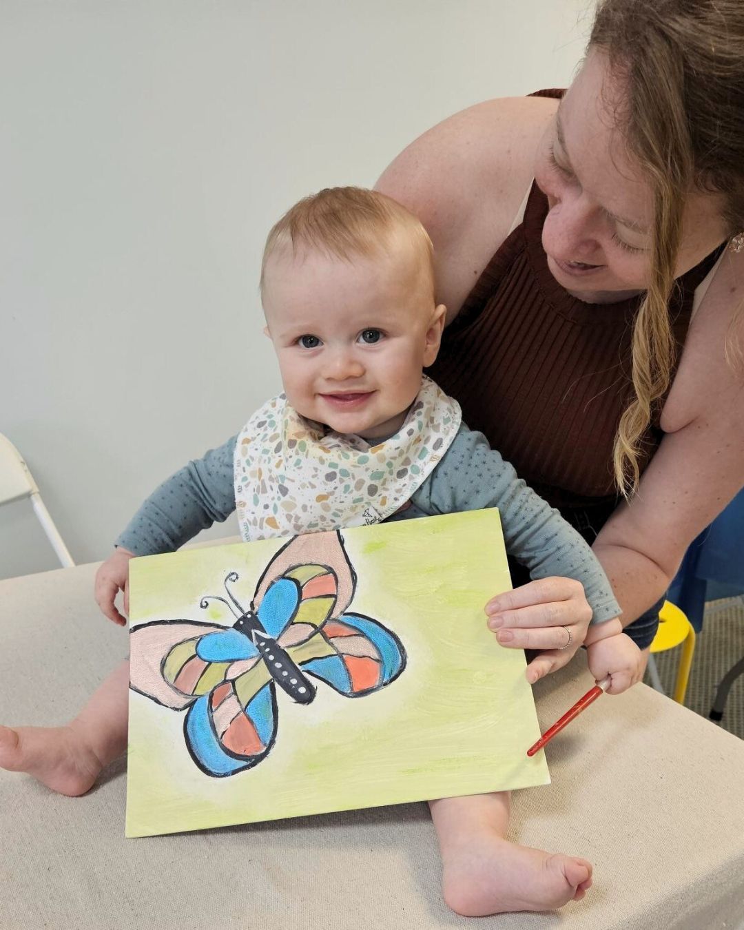 Baby holding a drawing of a butterfly with a woman sitting behind them.