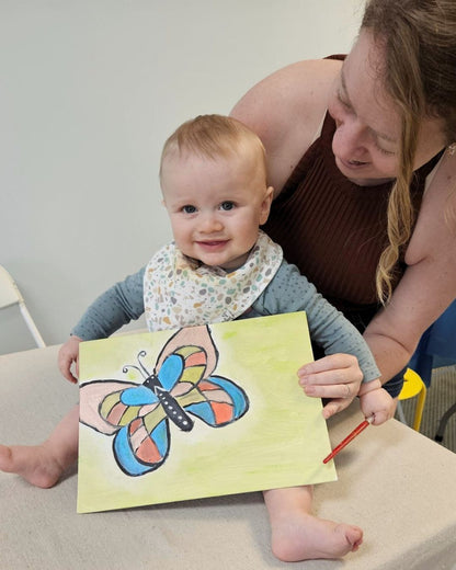 Baby holding a drawing of a butterfly with a woman sitting behind them.