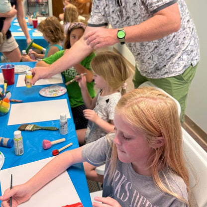 Man assisting a young girl with art supplies at a table in a classroom setting.