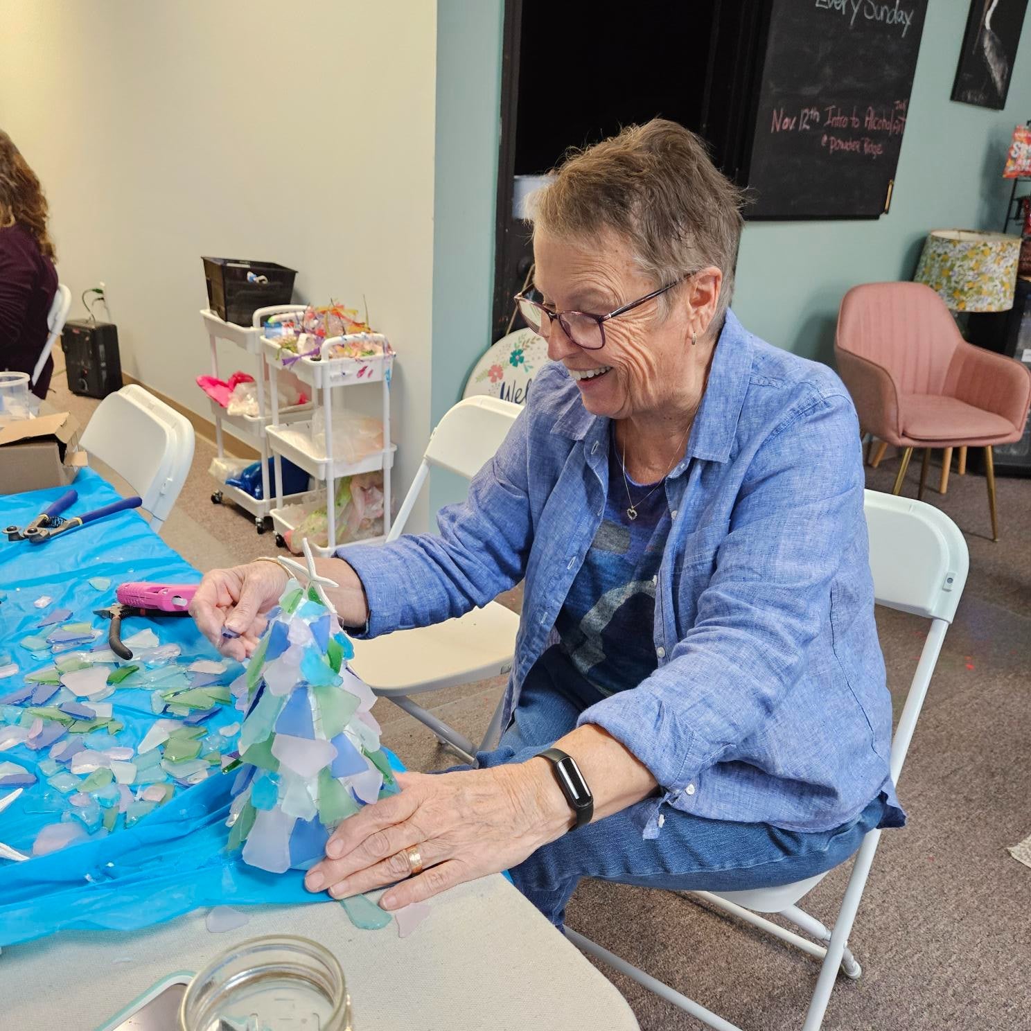 Woman crafting with sea glass at a table in a room with chairs and a chalkboard.